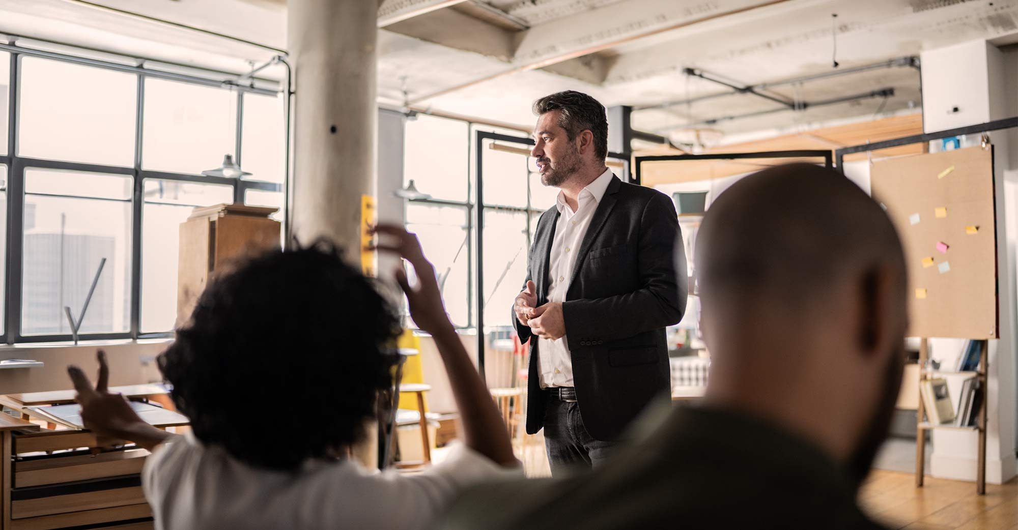 A man in a suit stands and speaks in a bright office while two people seated in the foreground, one raising a hand, listen and engage with him.