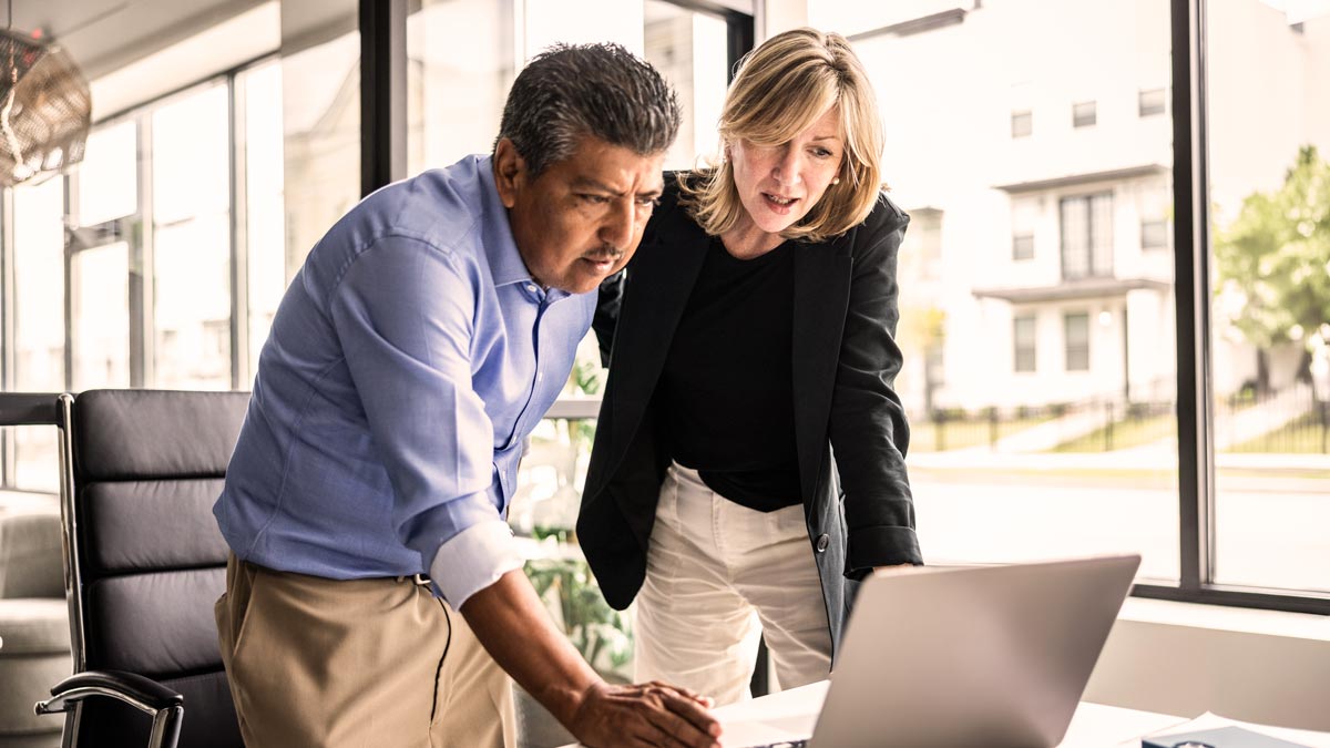 Two business professionals, a man and a woman, stand closely together in a modern office, looking intently at a laptop screen on a desk in front of them, with large windows and buildings visible outside.