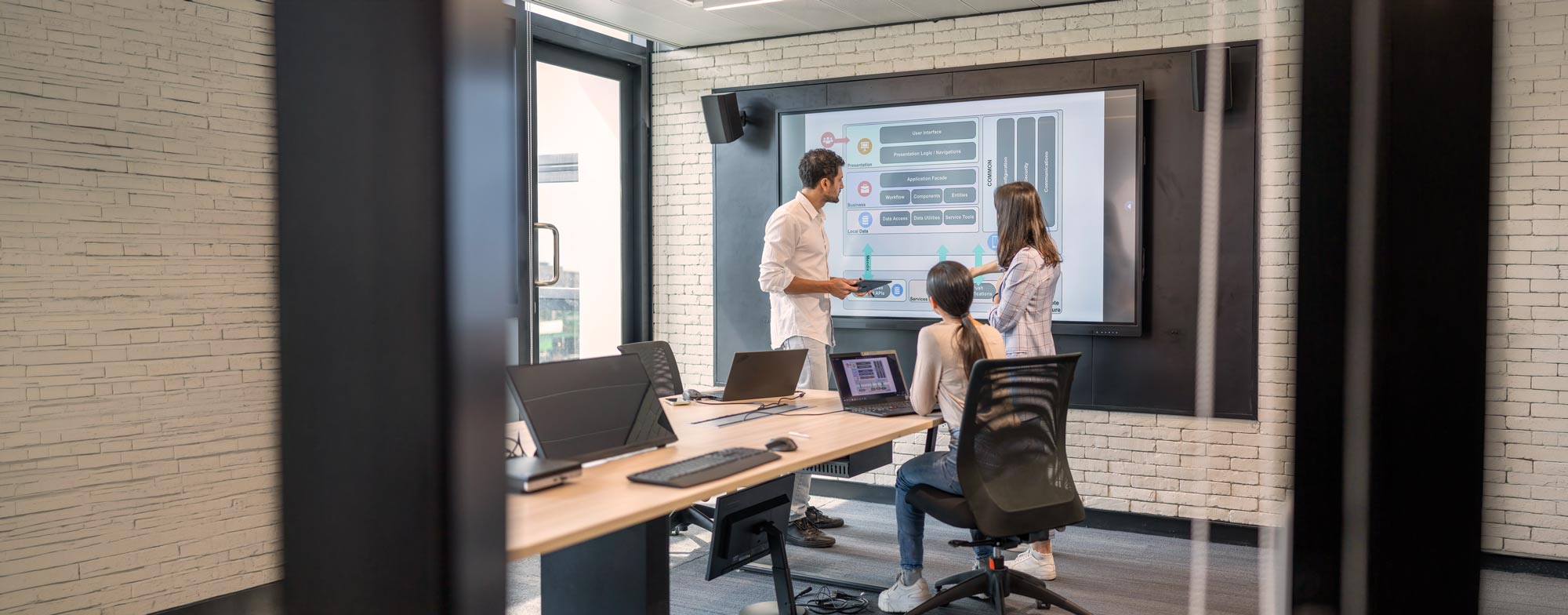 Three people in a modern office discuss a flowchart displayed on a large screen. Two stand and point at the chart, while one sits at a desk with open laptops. The room has glass walls and brick accents.