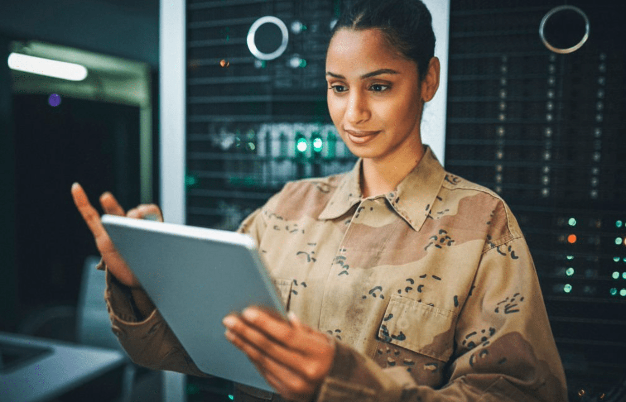 A woman in camouflage uniform uses a tablet while standing in a server room with computer equipment and glowing green lights in the background.