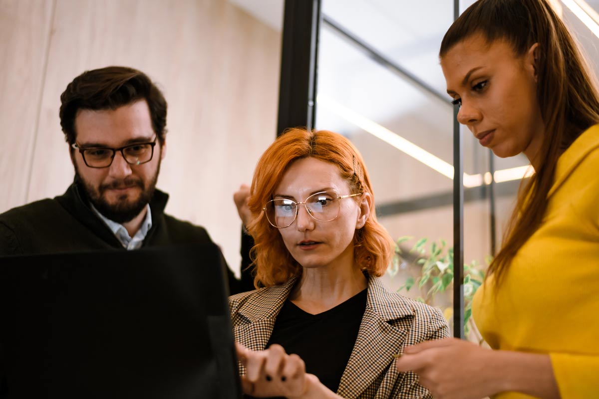Three people stand together in an office, focused on a laptop. One woman types while the others watch intently. The group appears to be collaborating or discussing something on the screen, possibly working on compliance automation strategies for their MSSP operations.