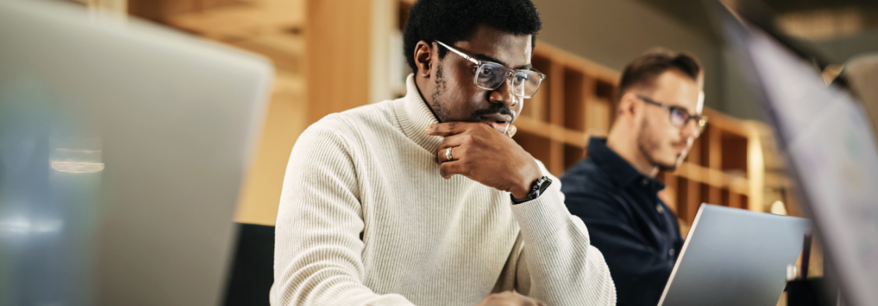Two men work at laptops in a modern office. One man in a white sweater and glasses is focused, resting his chin on his hand as he reviews data for a SOC 2 audit. Shelves and warm lighting are visible in the background.