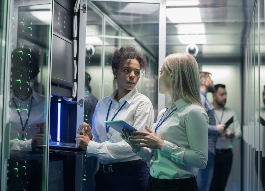 Two women in white shirts stand in a server room discussing work related to Certified Cloud Service Providers; one holds a tablet. Computer servers and two other people are visible in the background through glass panels, highlighting a modern environment focused on Rev5 and FedRAMP compliance.